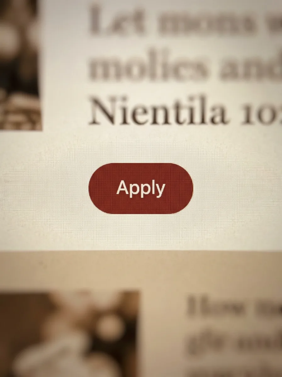 An extreme close-up of a brick-red pill-shaped Apply button on a cream homepage.